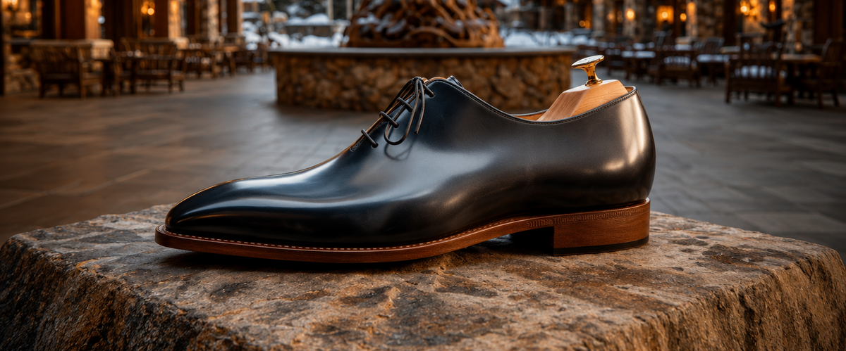 A black leather oxford dress shoe with a brown sole and heel, resting on a large stone pedestal in front of a blurred resort-style lodge courtyard with snow in the background.